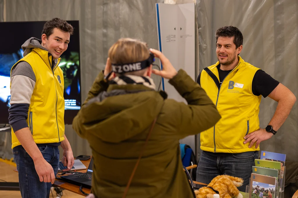 Visitatore con occhiali VR presso uno stand con due uomini in gilet giallo.